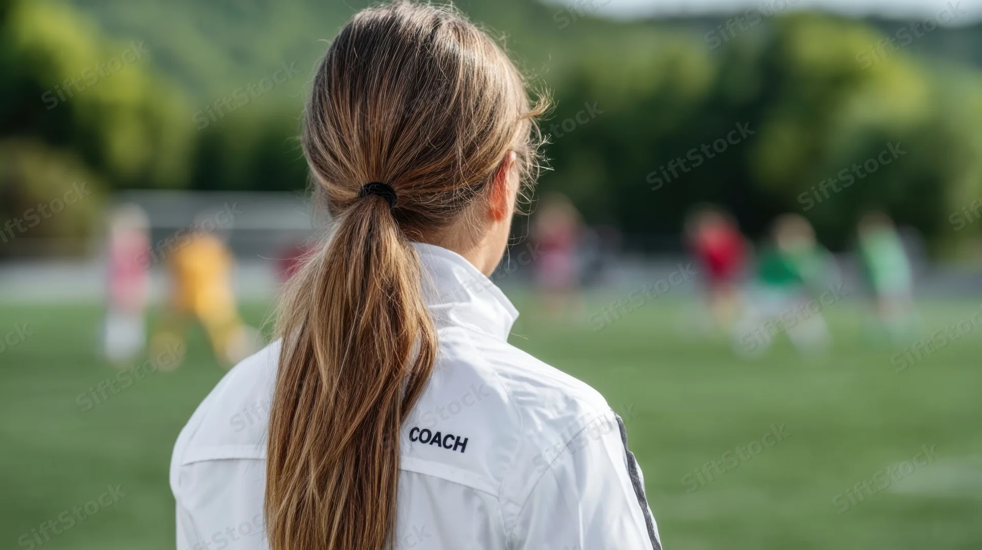 Woman coach in white jacket viewed from behind on soccer field with players in background