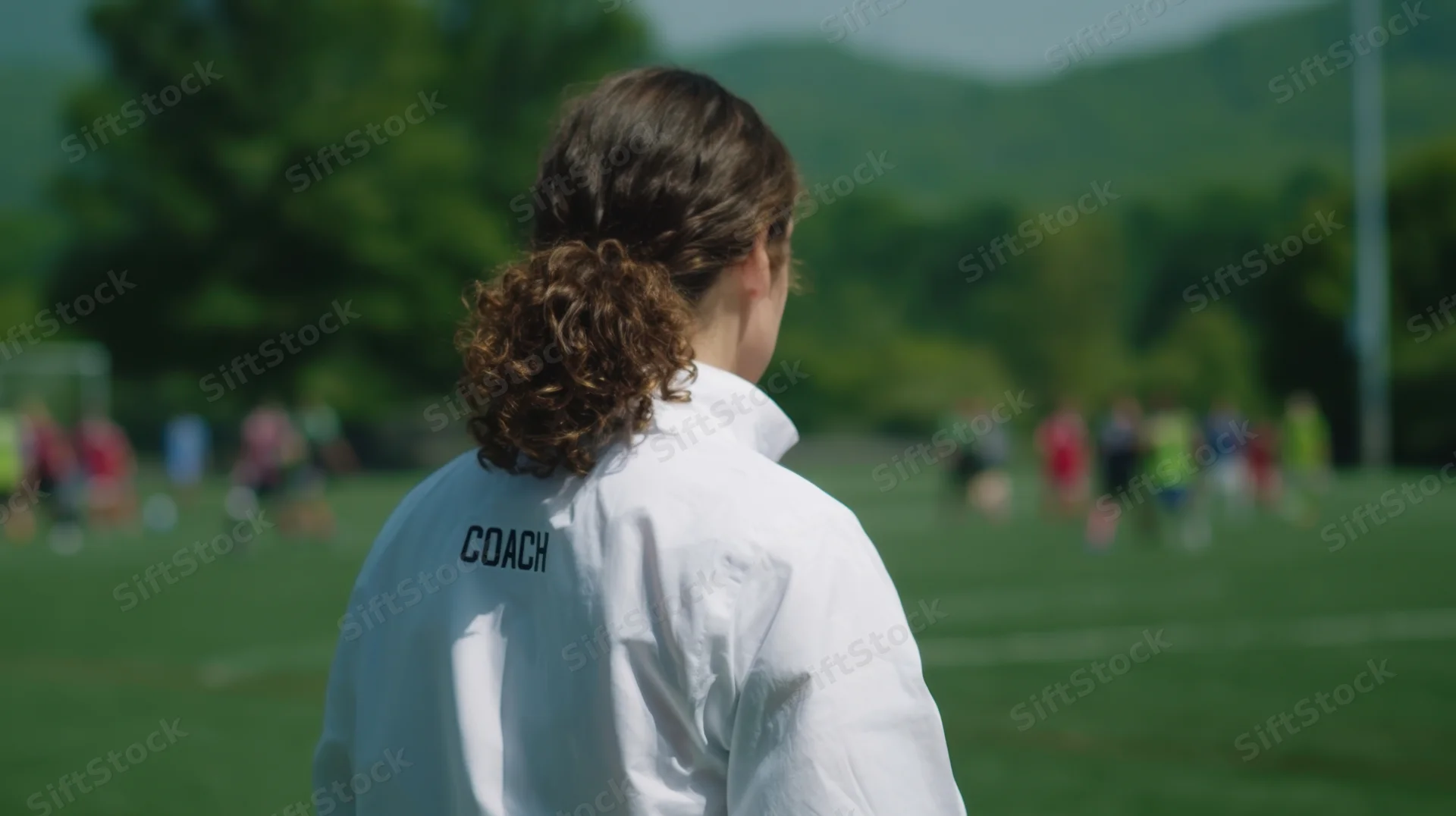 Woman in white coach jacket viewed from back on grass field with children playing soccer in background