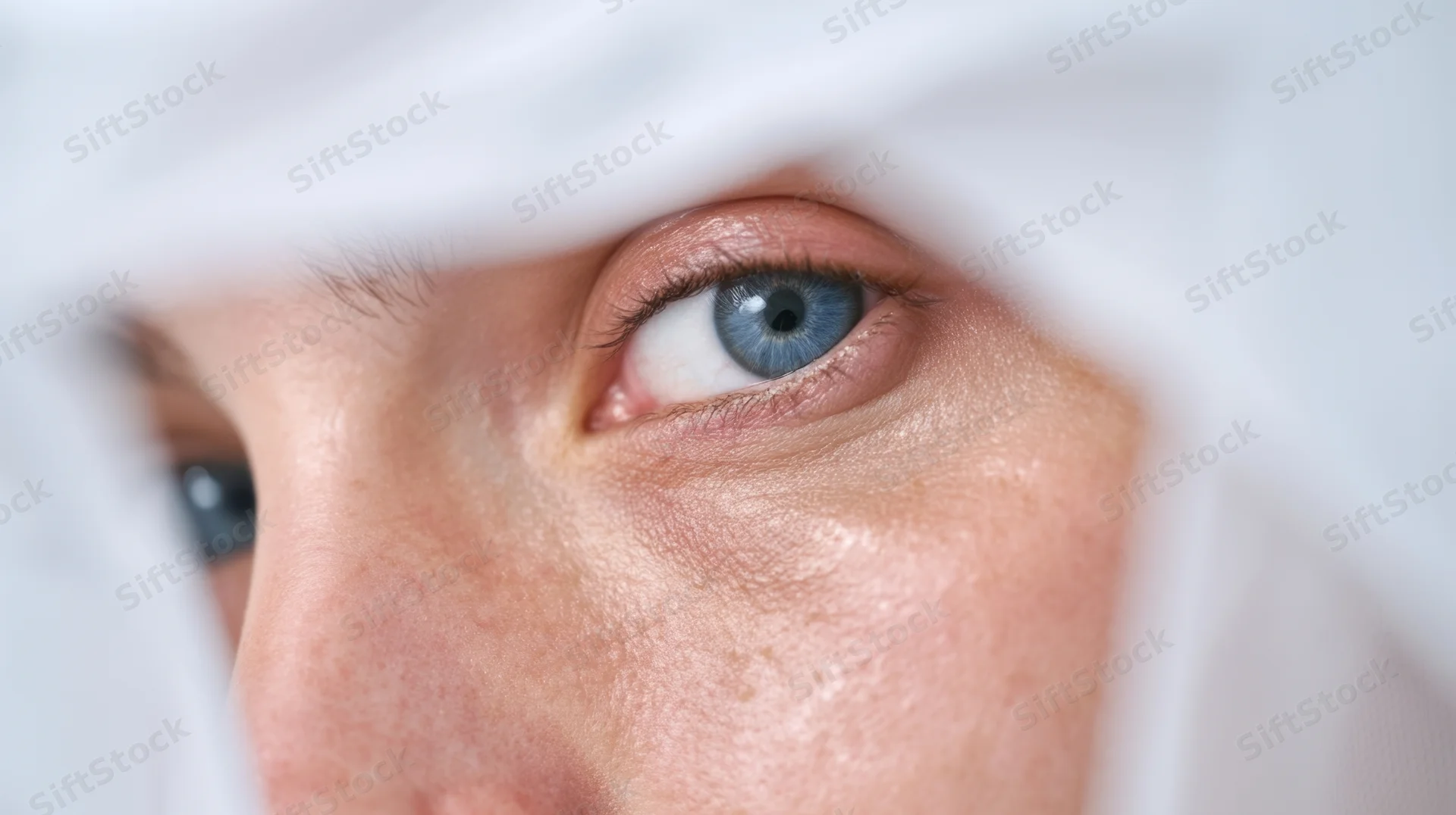 Close-up of a woman’s blue eye peeking through a white towel during a facial treatment