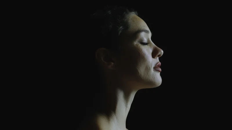 Woman in profile with closed eyes against dark background, illuminated by soft light on face and neck