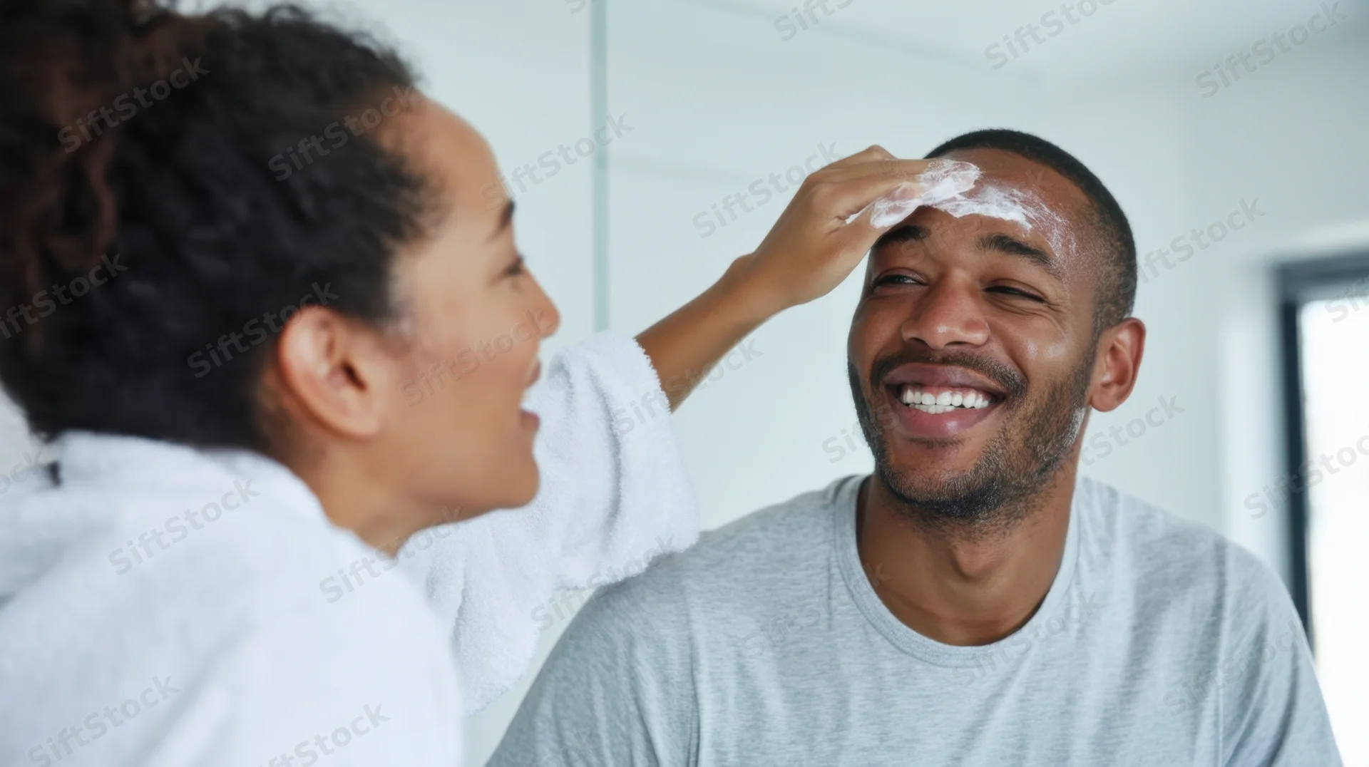 Woman applying moisturizer to man's forehead in bathroom mirror, both laughing
