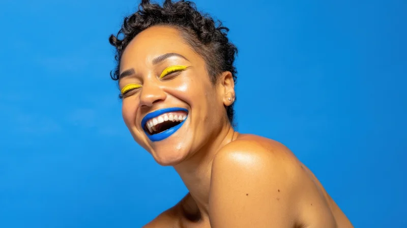Woman laughing with bright blue lipstick and yellow eyeliner against a blue background