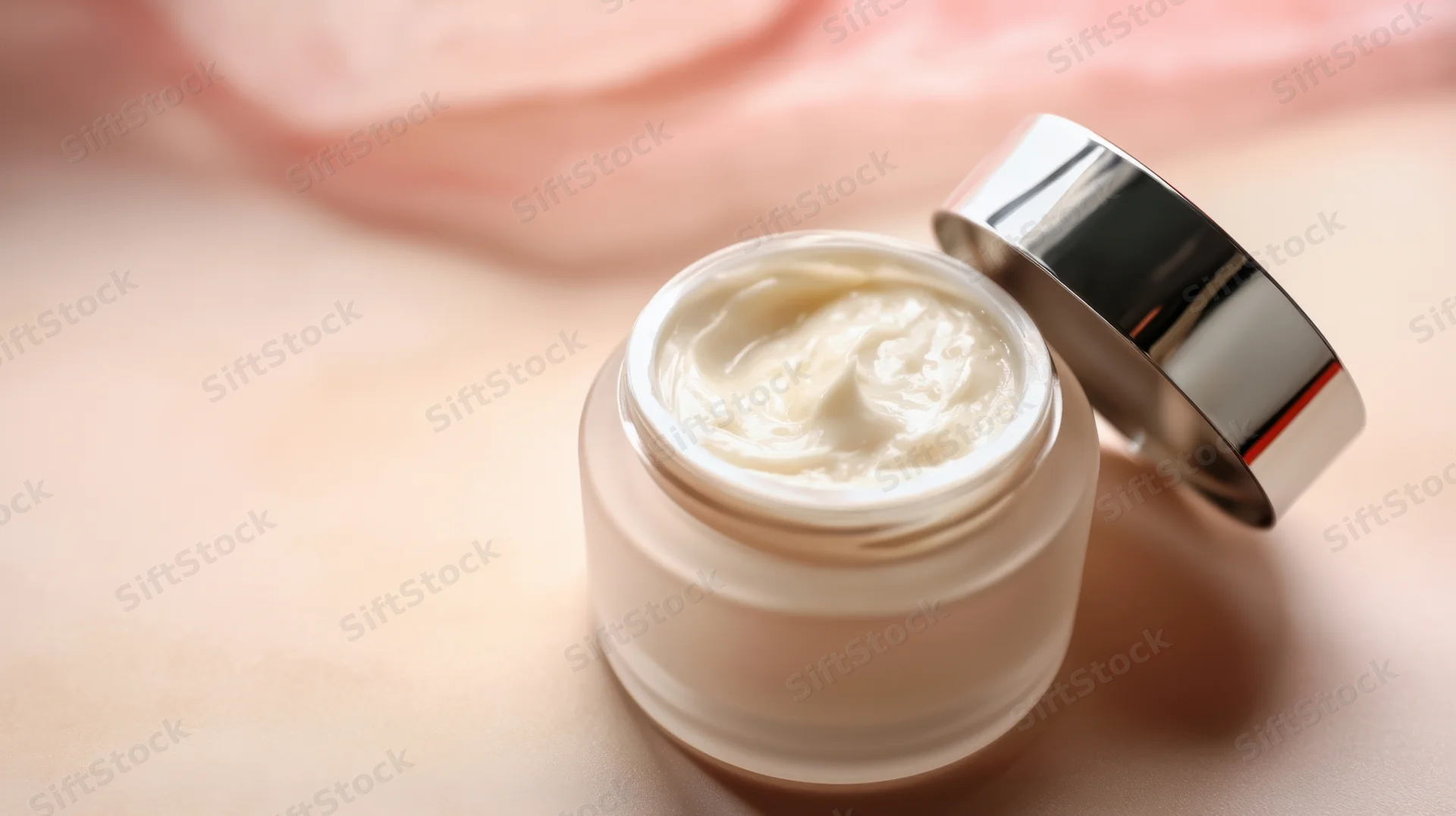Open jar of white cream on soft pink surface with lid beside it