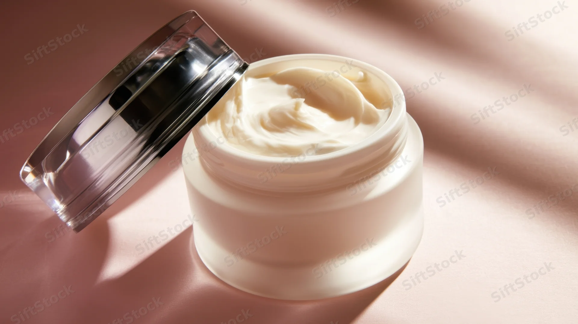 Open jar of white cream on pink satin surface with lid beside it