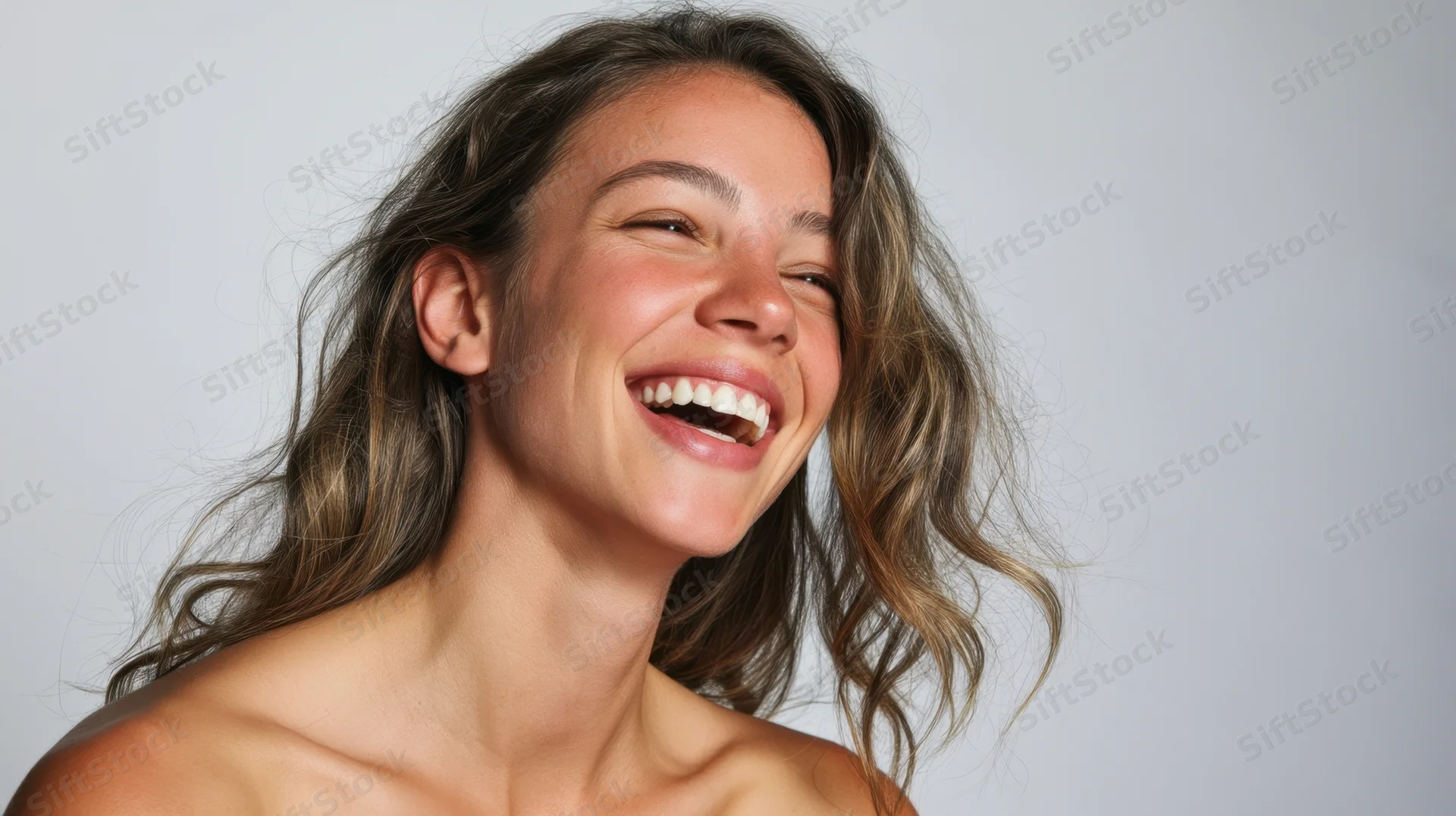 Young woman laughing with wavy brown hair, bare shoulders, against light gray background