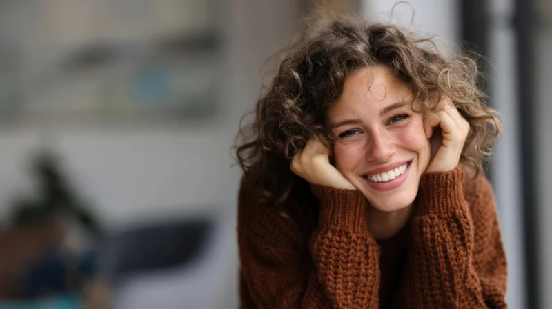 Young woman smiling with hands on cheeks, wearing brown knit sweater indoors