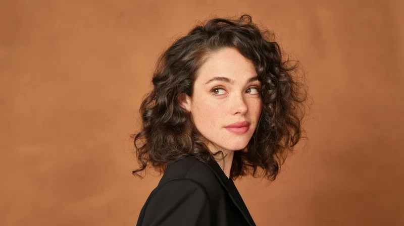 Young woman with curly brown hair in black blazer turning head in studio portrait against terracotta background