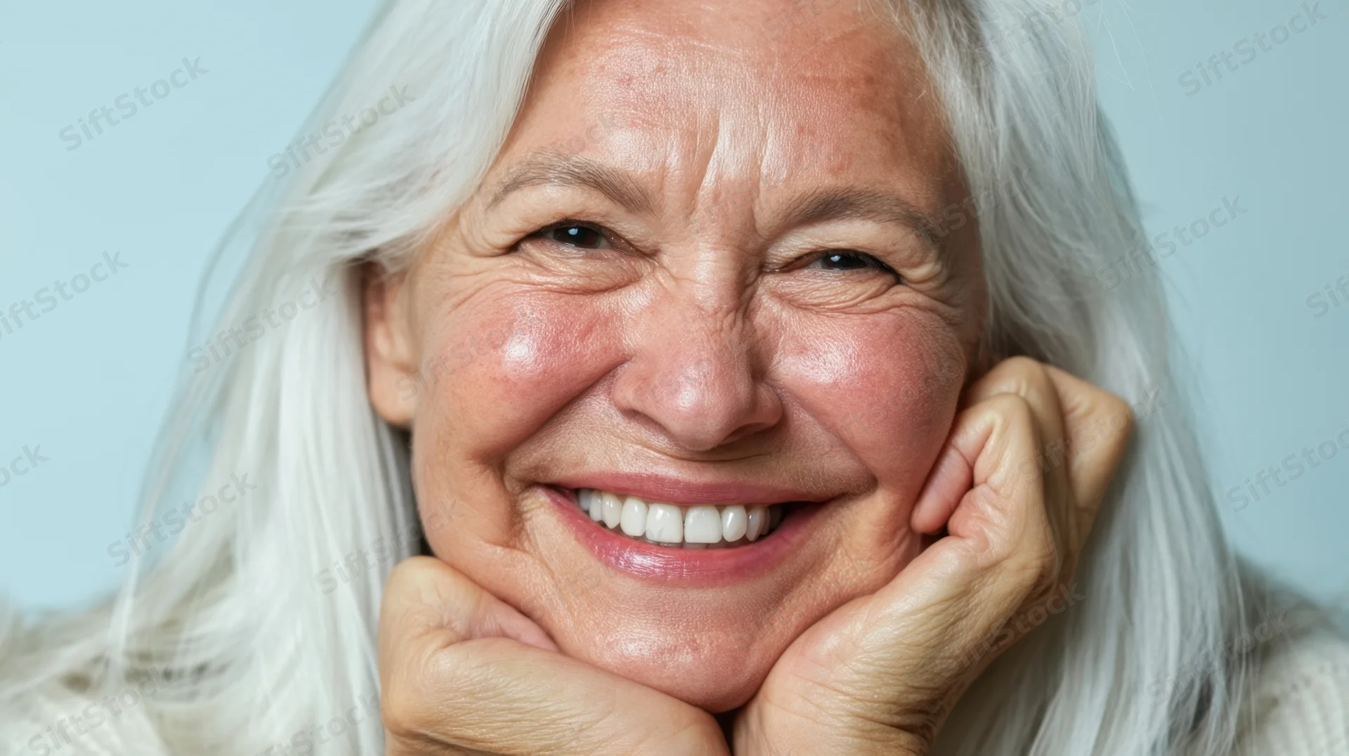 Smiling senior woman with white hair resting her chin on her hands against a light blue background