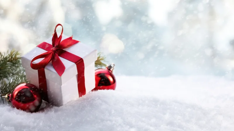 White gift box with red ribbon and red ornaments in snow near pine branches