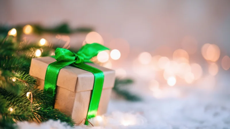 Gift box wrapped in brown paper with green ribbon resting on snowy surface beside pine branches and glowing lights