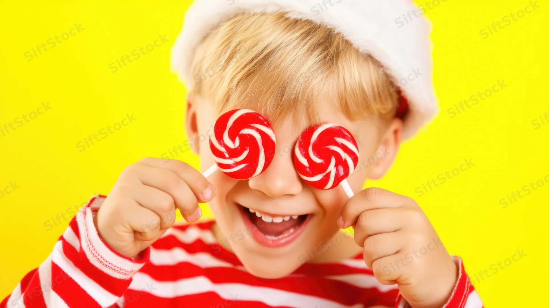 Boy holding lollipops over eyes while wearing santa hat and striped sweater against yellow background