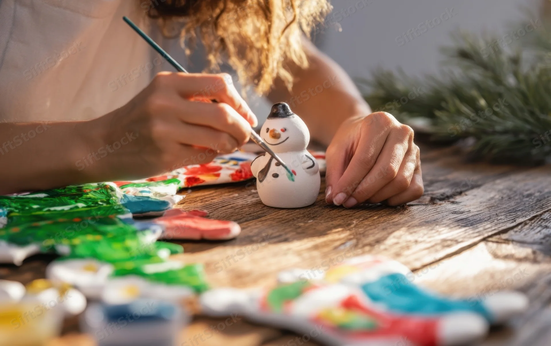 Woman painting a ceramic snowman decoration on a wooden table with craft supplies around