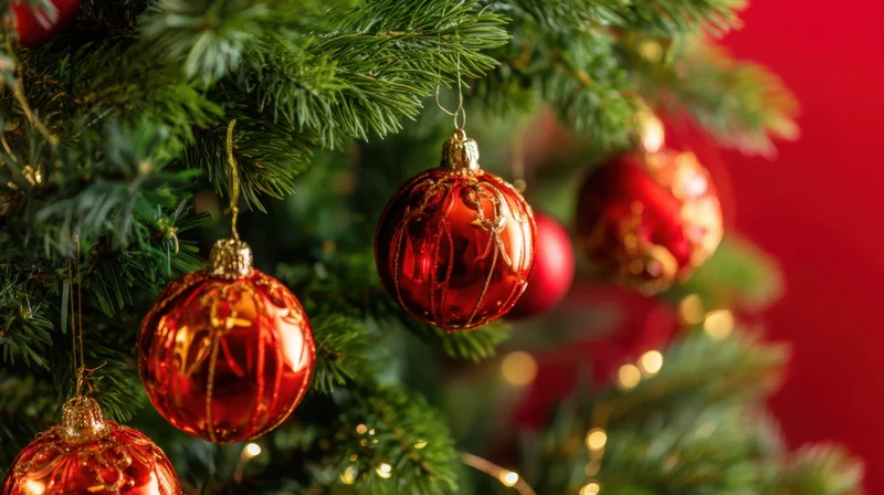 Red glass ornaments hanging on a christmas tree branch with warm lights and a red background
