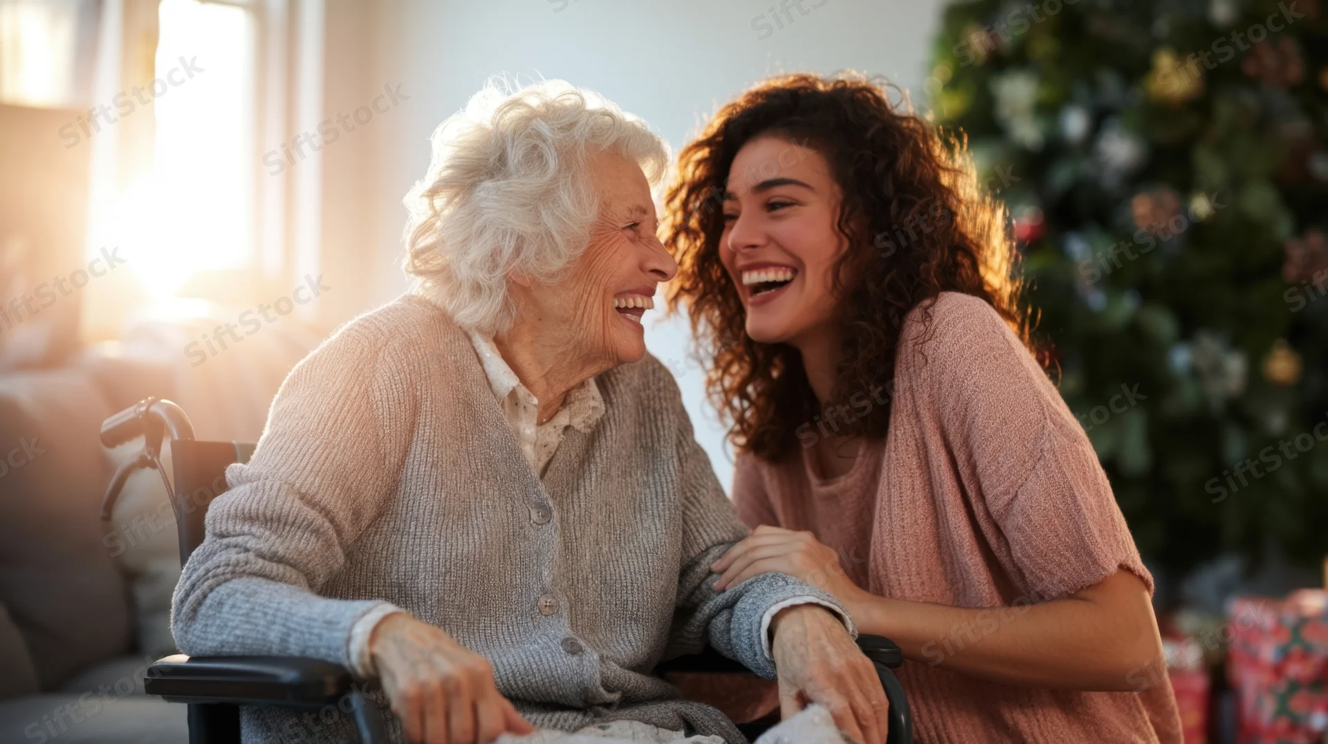Young woman laughing with senior woman in wheelchair by christmas tree at home
