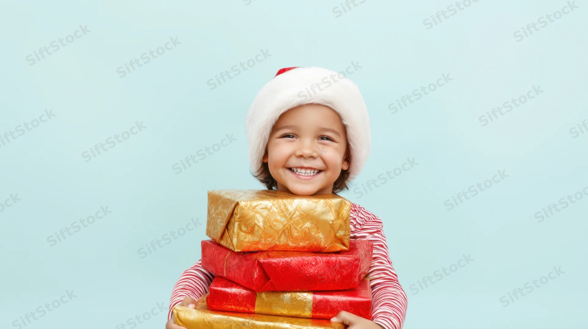 Child smiling while holding a stack of wrapped gifts, wearing a santa hat against a light blue background