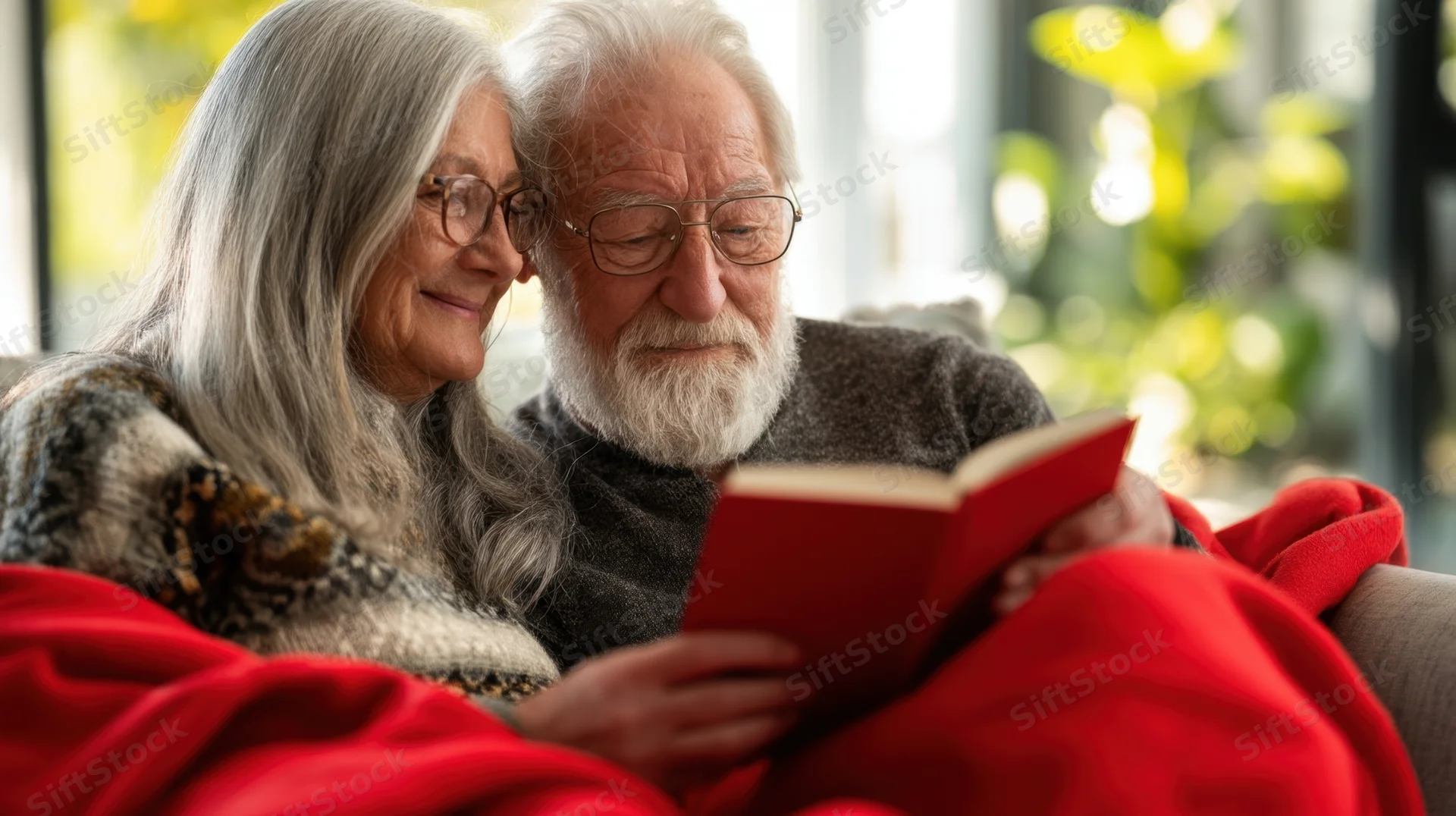 Elderly couple reading a book together under a red blanket in a cozy living room
