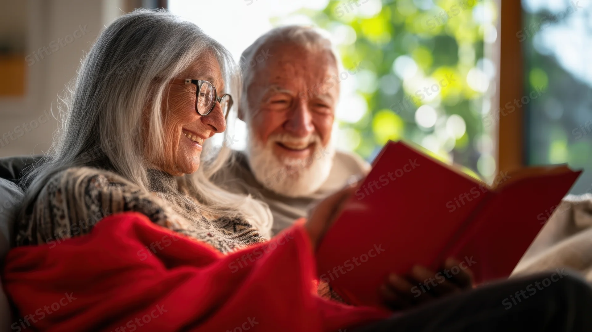 Elderly couple smiling while reading a book together on a sofa at home