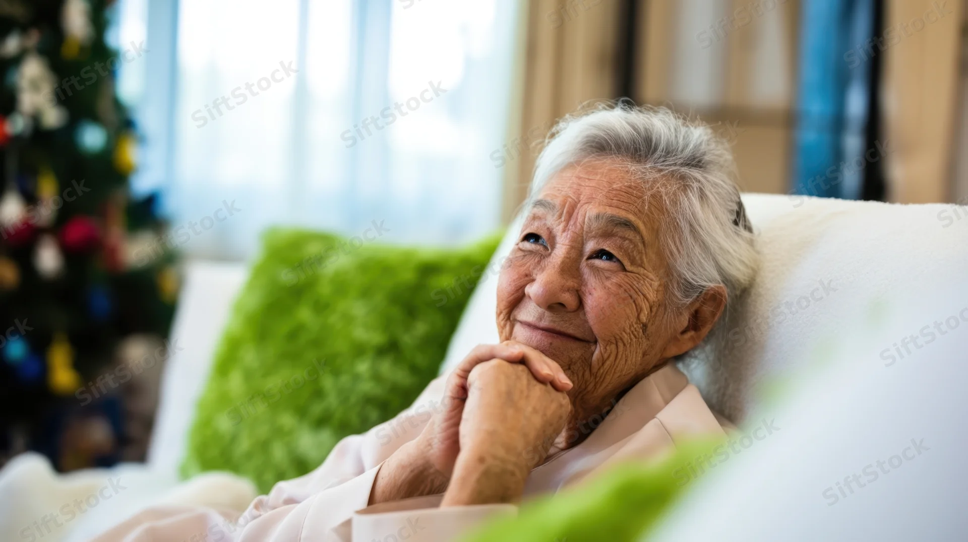 Senior asian woman smiling with hands clasped while relaxing on a couch near a decorated christmas tree in a living room