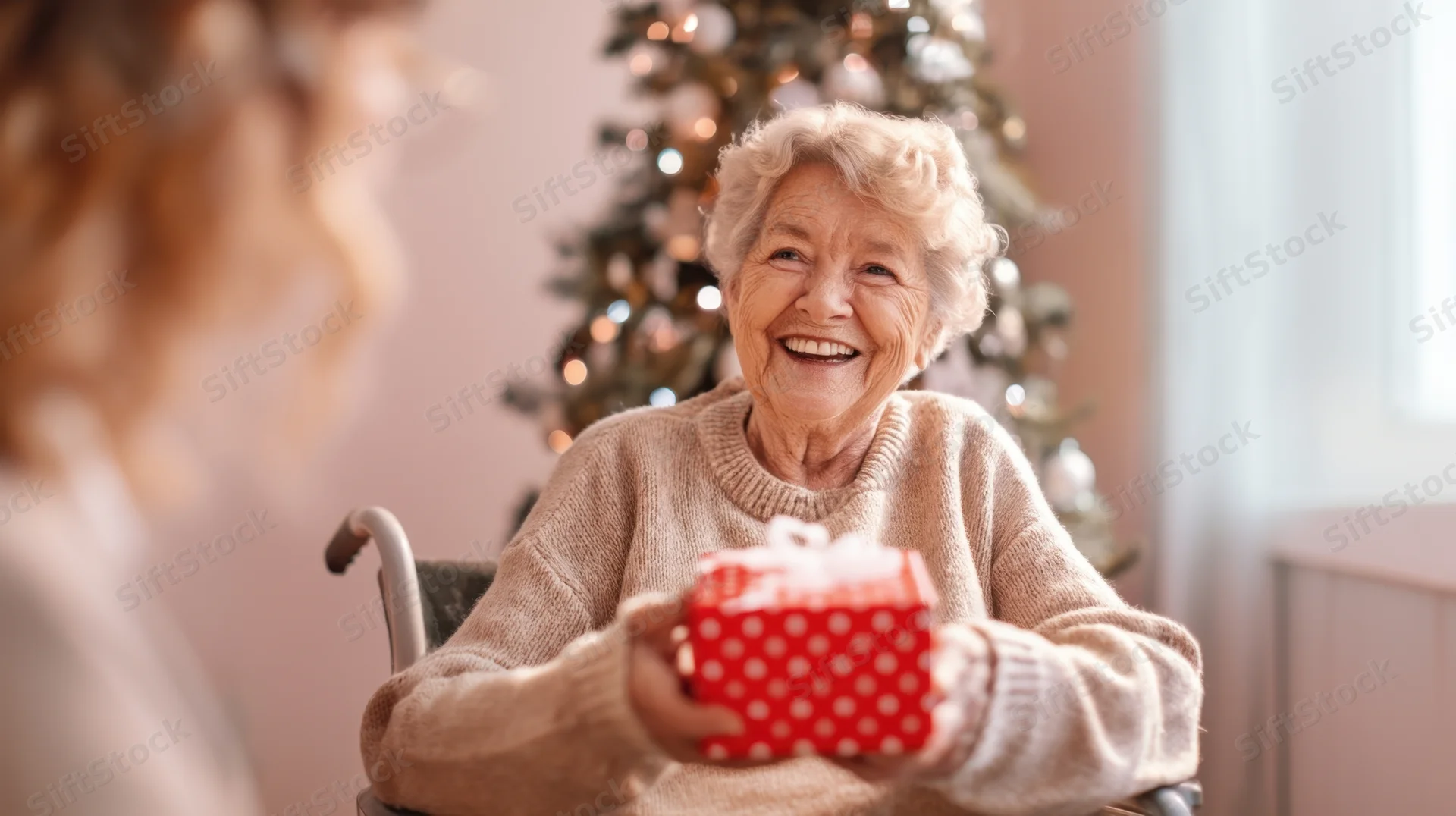 Senior woman smiling while holding a red polka dot gift box in a wheelchair near a decorated christmas tree indoors