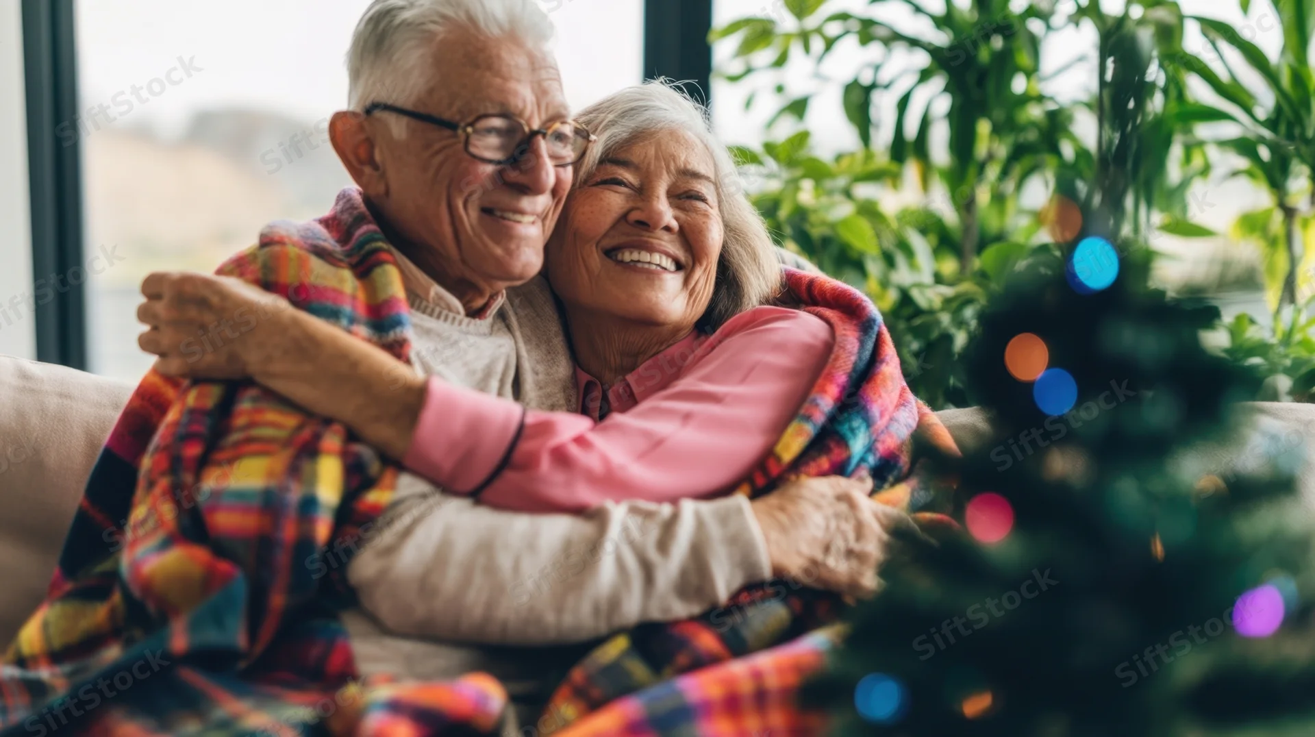 Elderly couple hugging on a couch wrapped in a plaid blanket near a christmas tree