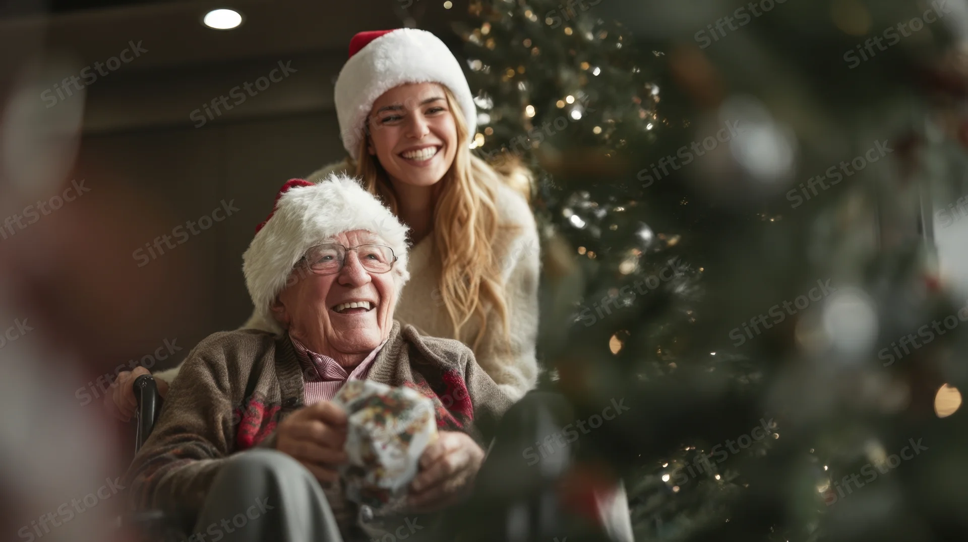 Elderly man in wheelchair smiling while holding a gift, with a young woman behind him, both wearing santa hats near a decorated christmas tree indoors