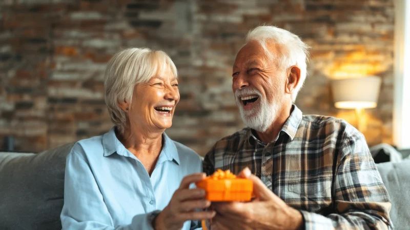 Elderly couple laughing while holding a small orange gift box on a sofa in a cozy living room