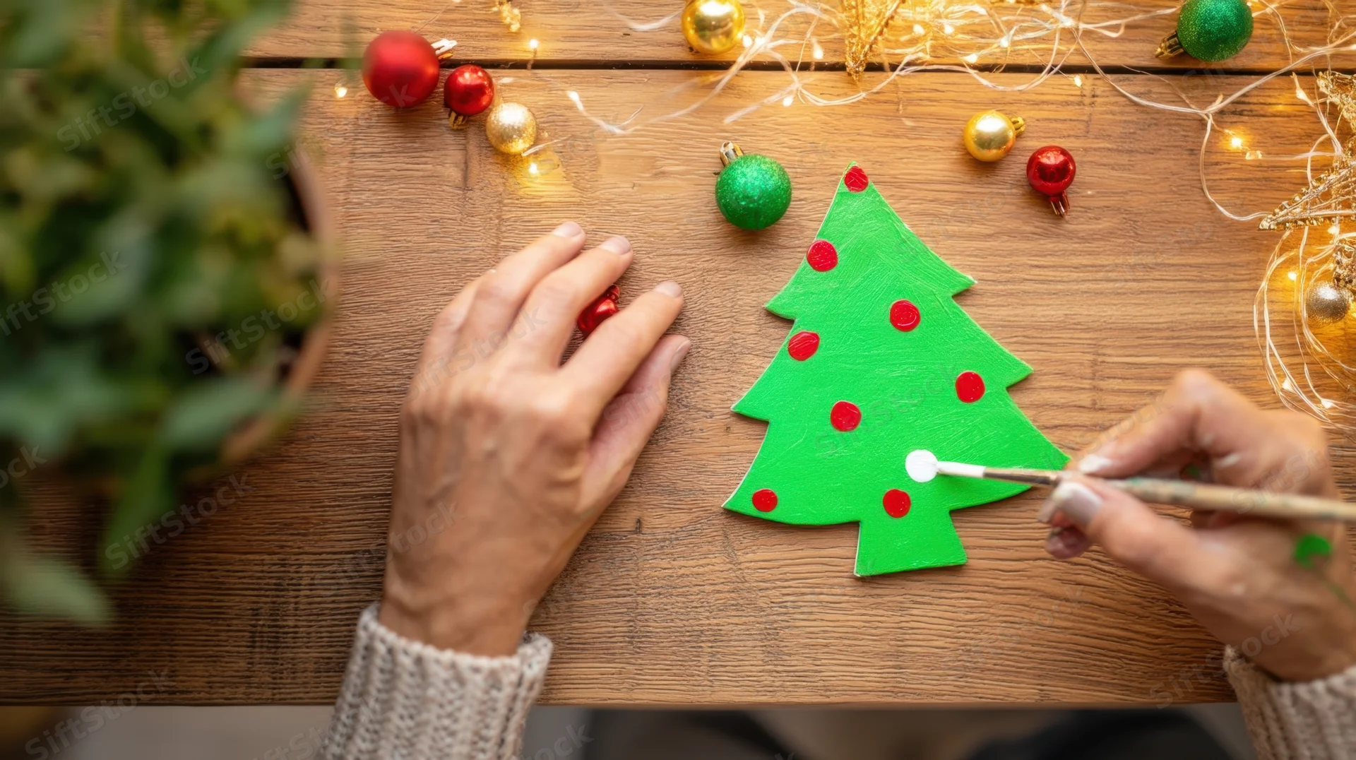 Woman painting a green christmas tree cutout with red dots on a wooden table surrounded by ornaments and fairy lights