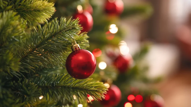 Red shiny ornament hanging on a green christmas tree branch with blurred lights in the background