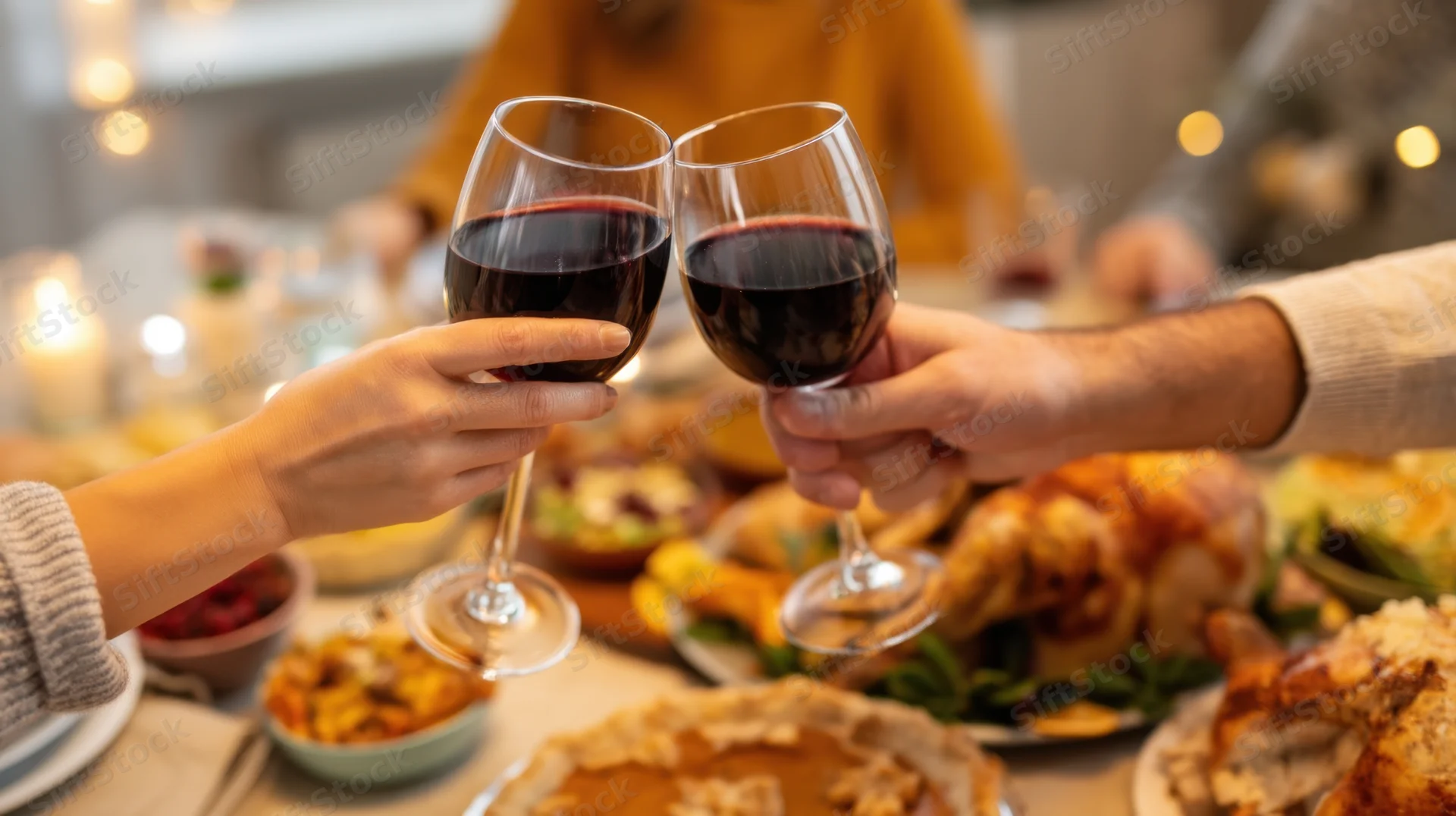 Two hands clinking wine glasses over a dinner table filled with turkey, pie and candles