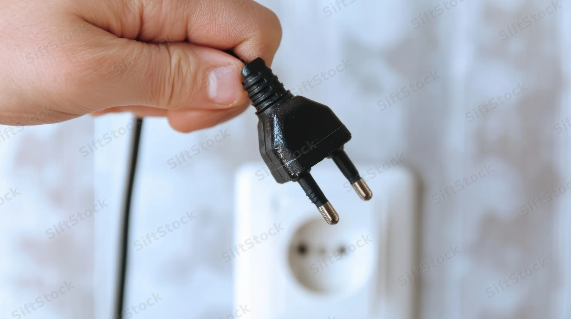 Hand holding black two-prong electrical plug near white wall socket