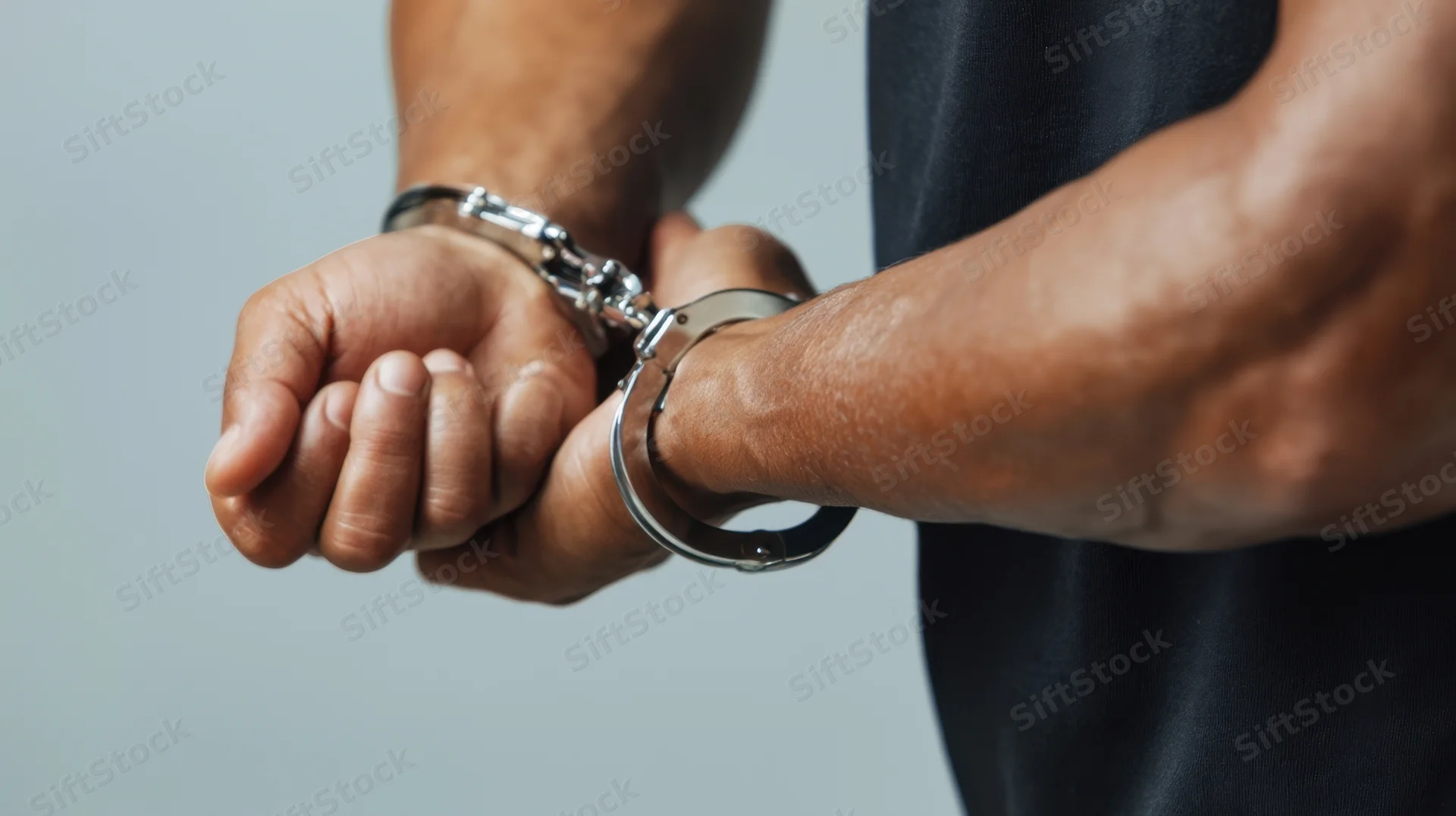 Man's hands cuffed behind his back in silver handcuffs, wearing black tshirt, studio shot
