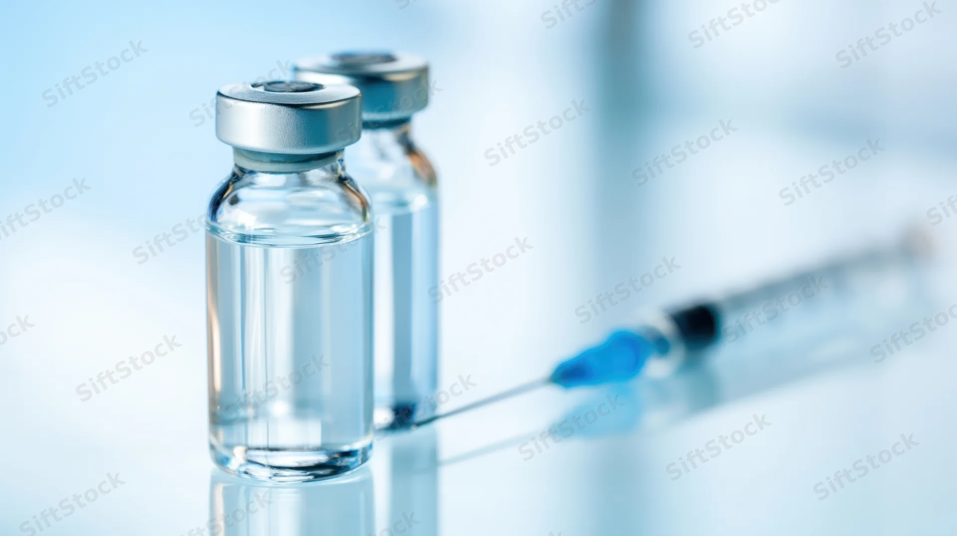 Two glass vaccine vials with a syringe on a reflective surface in a clinical setting