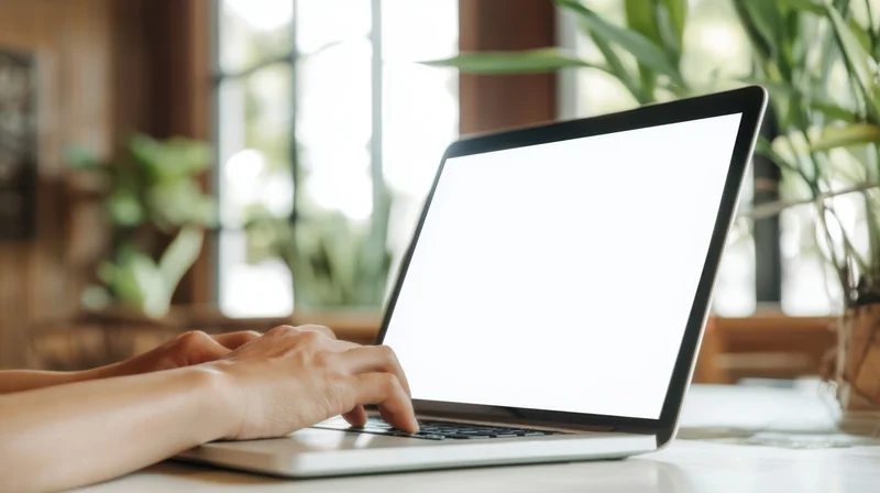 Hands typing on a laptop with blank white screen on a desk near a window with plants