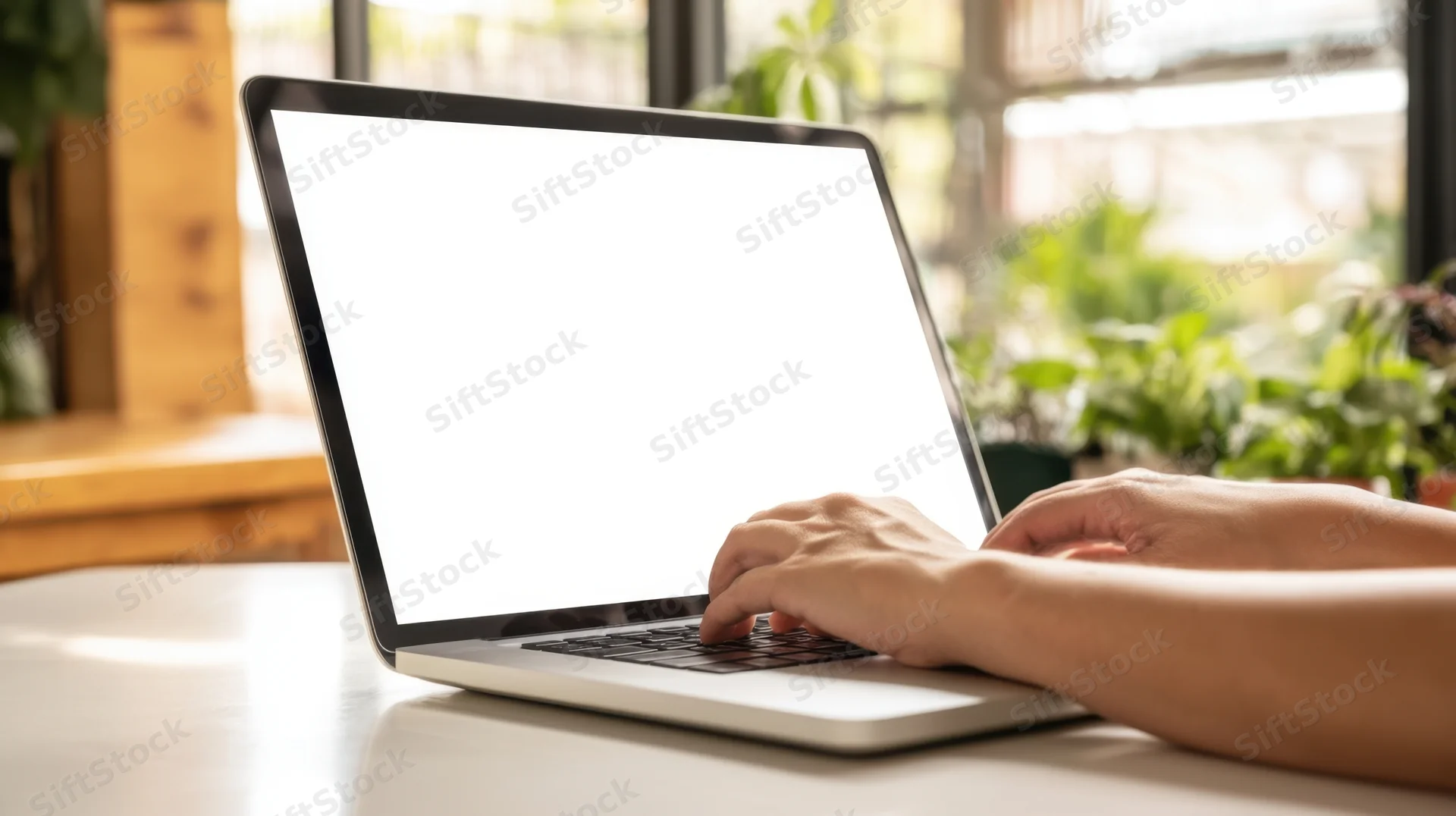 Hands typing on a laptop with blank white screen on a desk near a window with plants