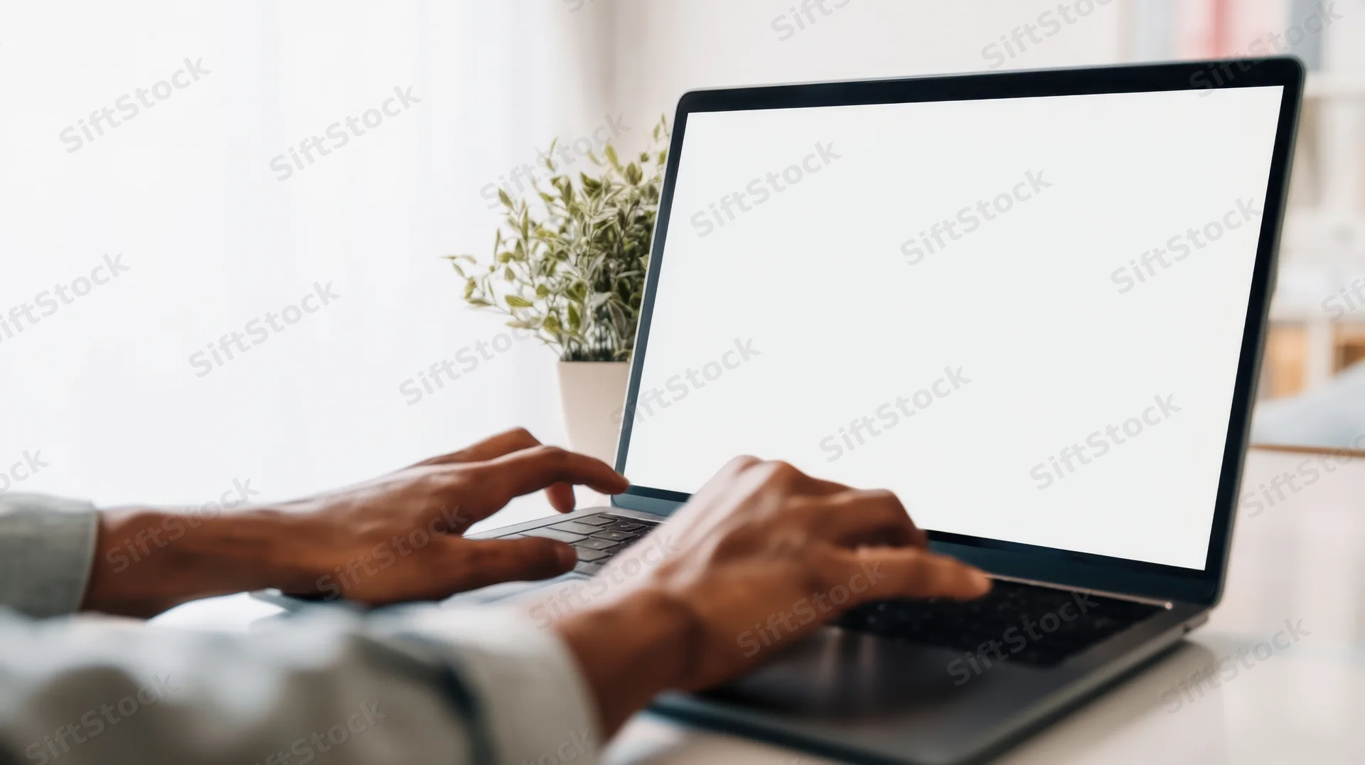 Hands typing on a laptop with blank white screen on a desk near a window