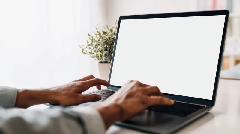 Hands typing on a laptop with blank white screen on a desk near a window