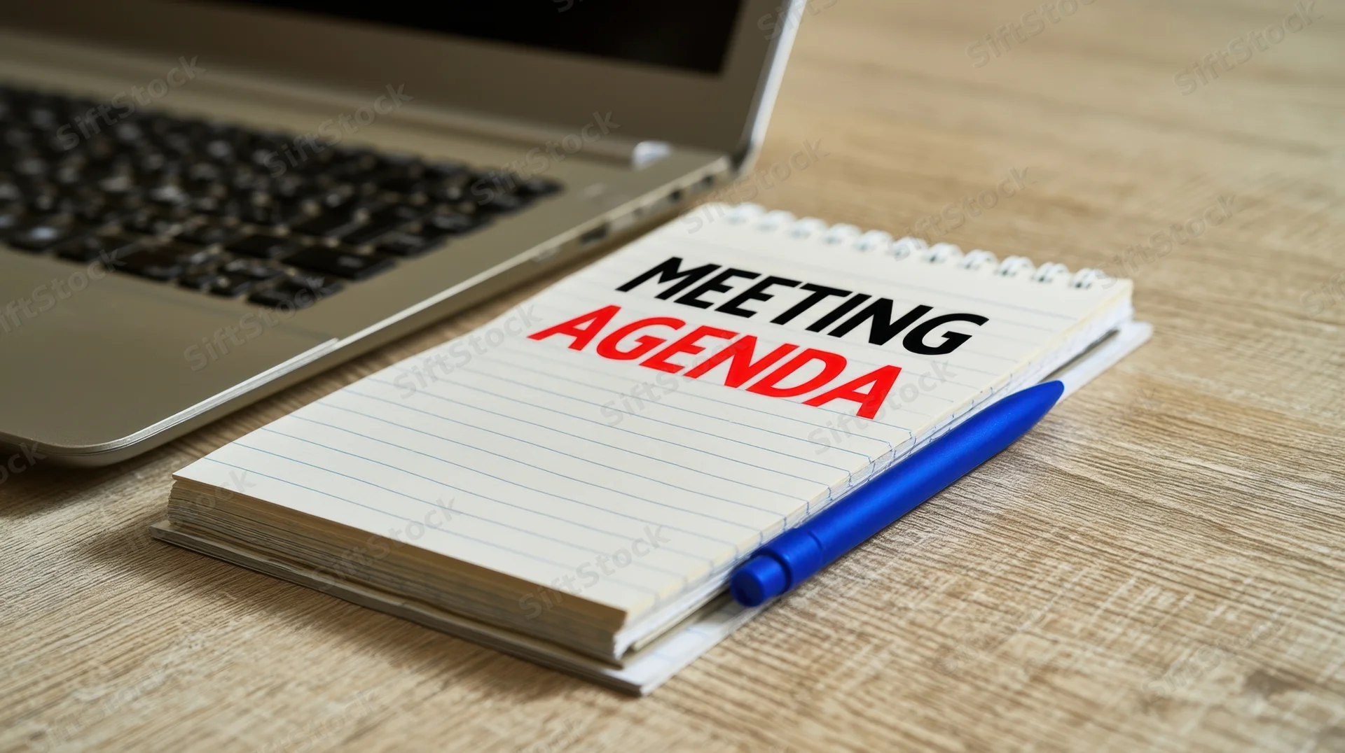 Notepad with meeting agenda text and blue pen on wooden desk beside laptop