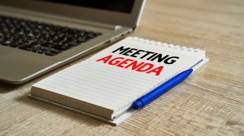Notepad with meeting agenda text and blue pen on wooden desk beside laptop