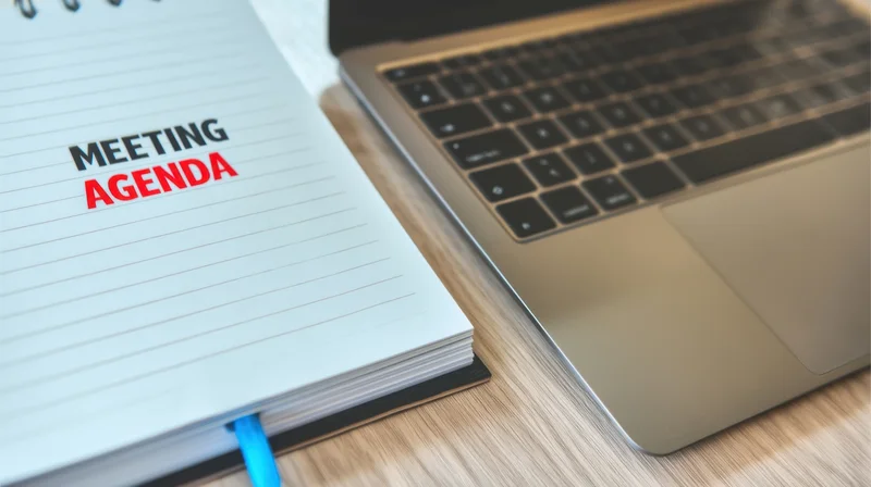 Notepad with meeting agenda text beside a laptop on a wooden desk