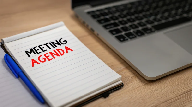 Notepad with meeting agenda written on it next to a laptop on a wooden desk