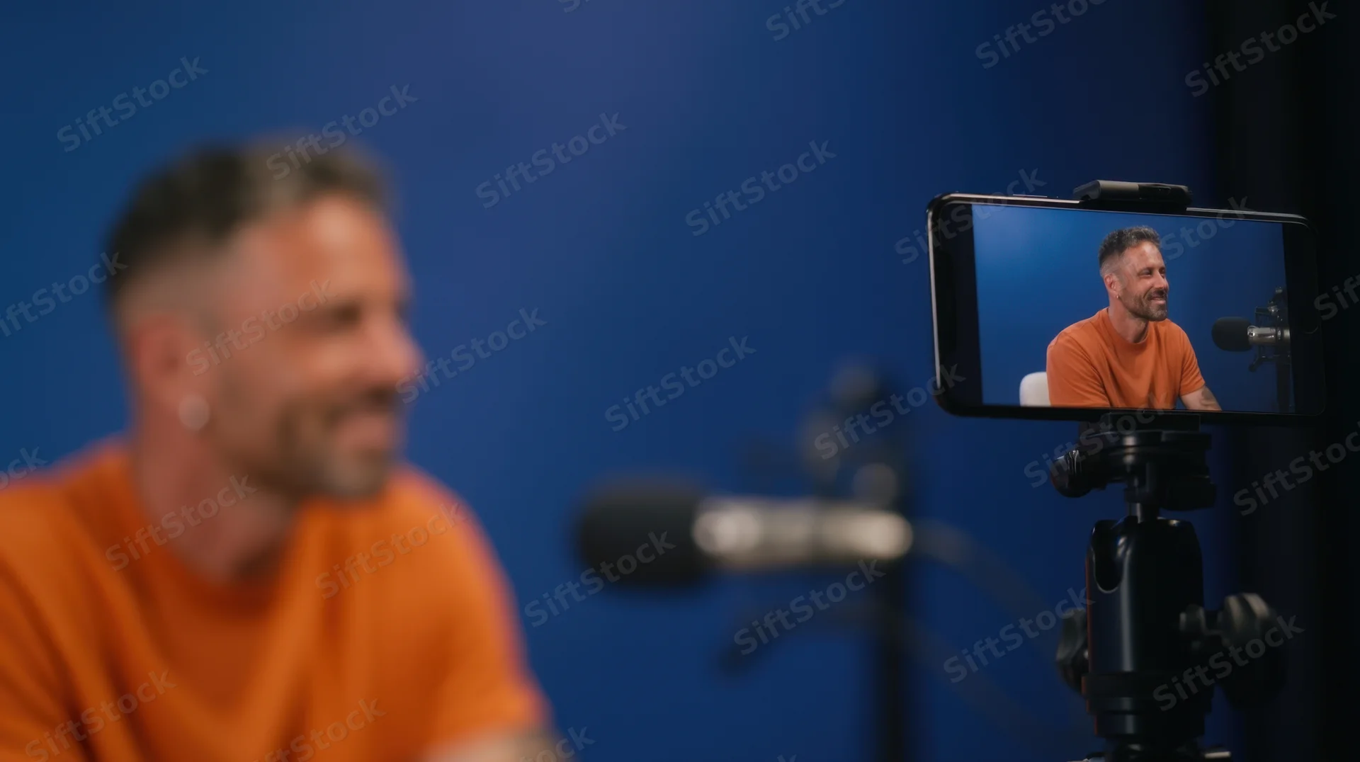 Man recording video content on smartphone in studio with microphone and blue background