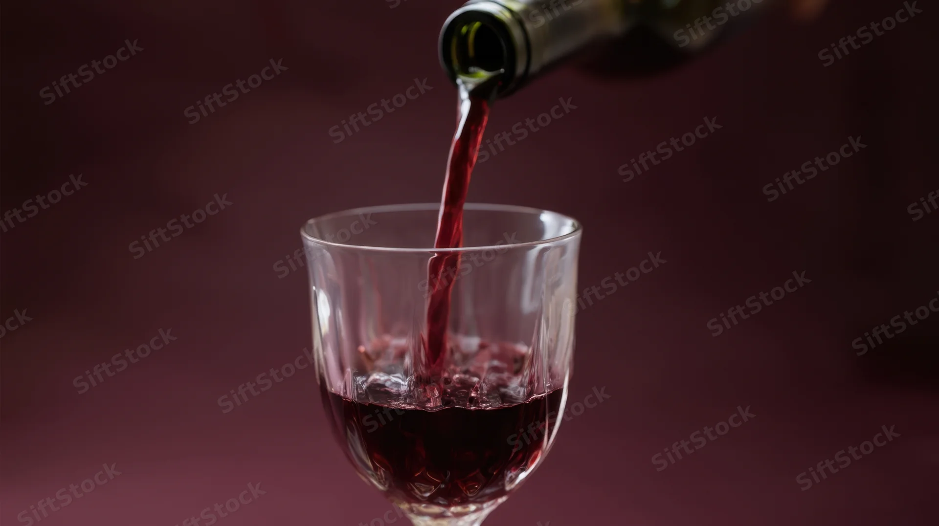 Red wine being poured from a bottle into a glass against a dark maroon background