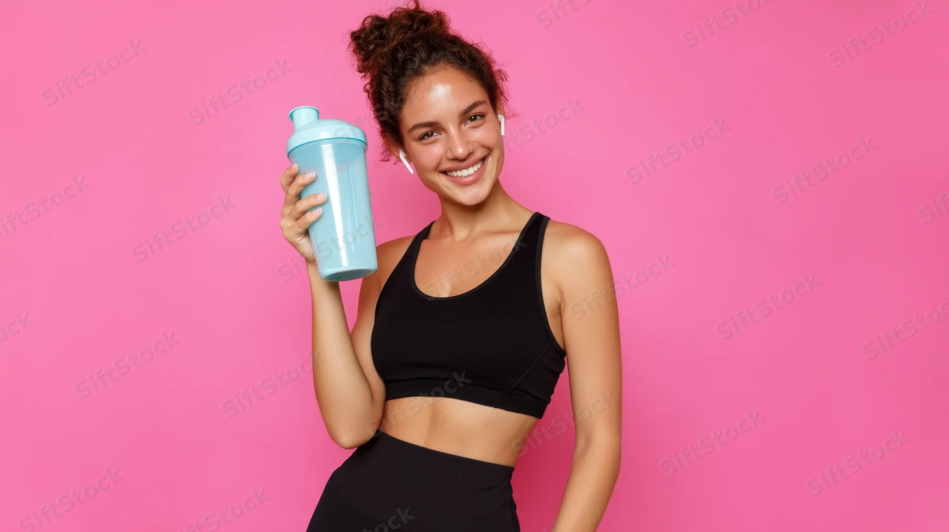 Young woman smiling while holding a blue shaker bottle against a pink background
