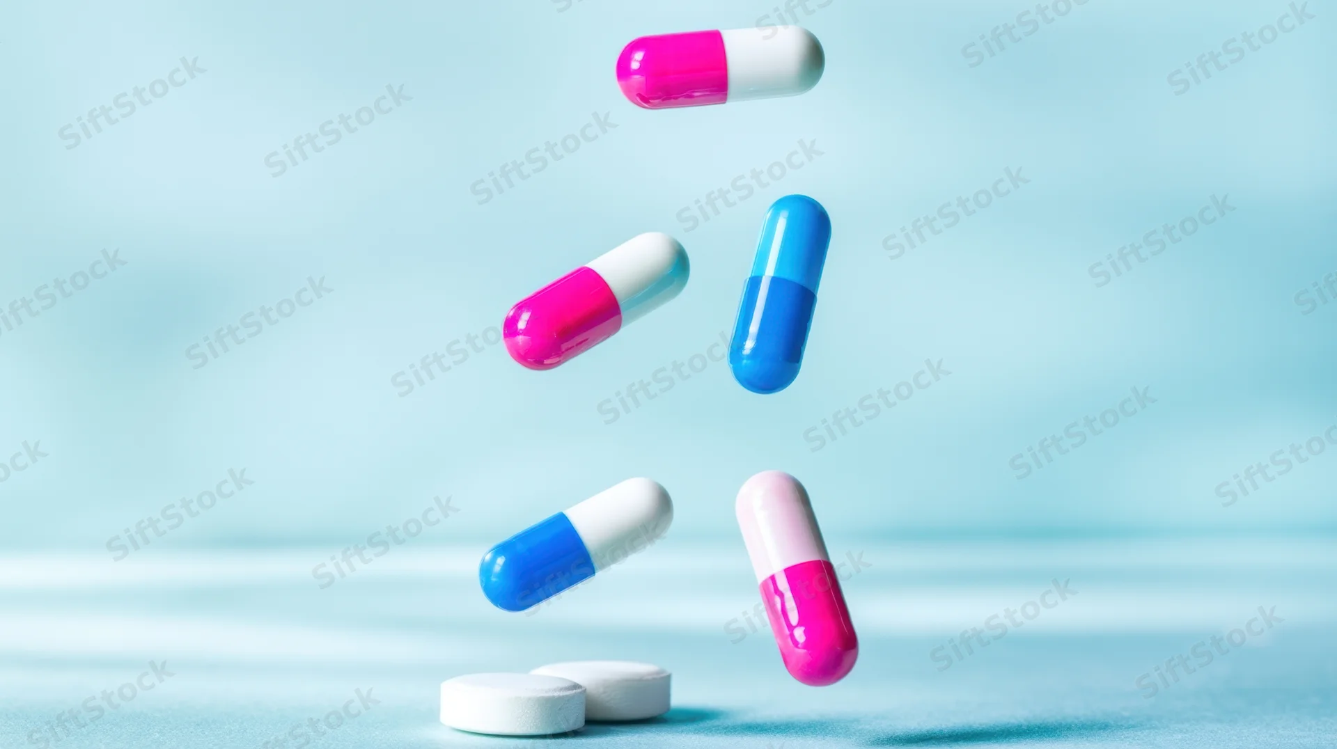 Colorful capsules falling midair onto a light blue surface with tablets below