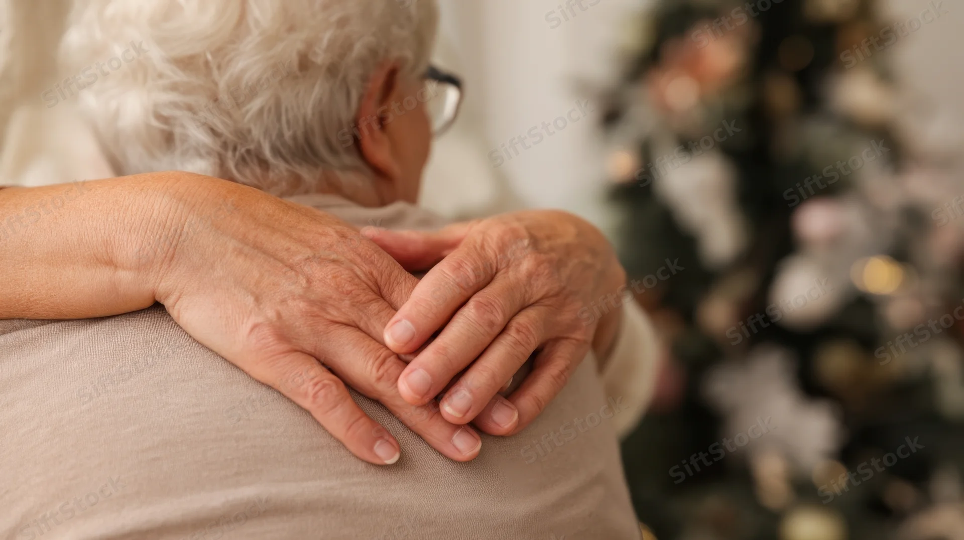 Senior woman seen from back receiving a comforting hug with hands on her shoulders near a decorated christmas tree