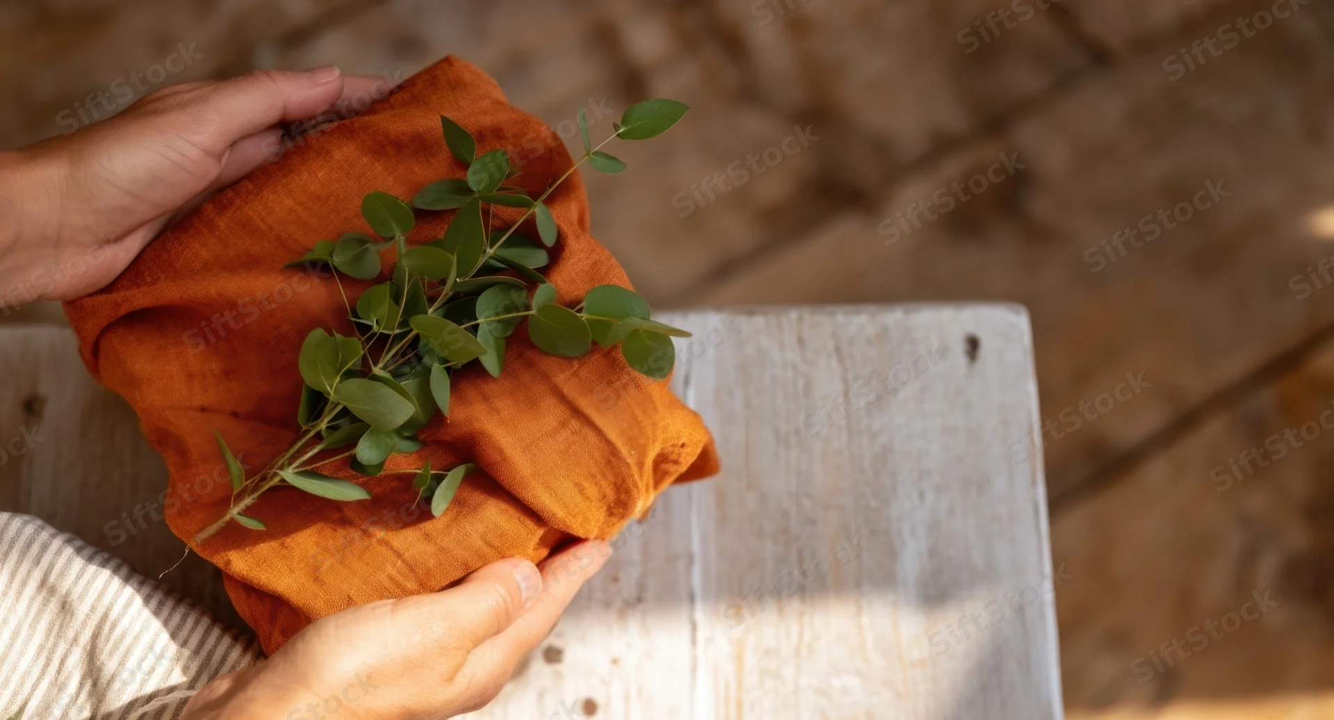 Hands holding a cloth-wrapped gift with eucalyptus leaves on a wooden table