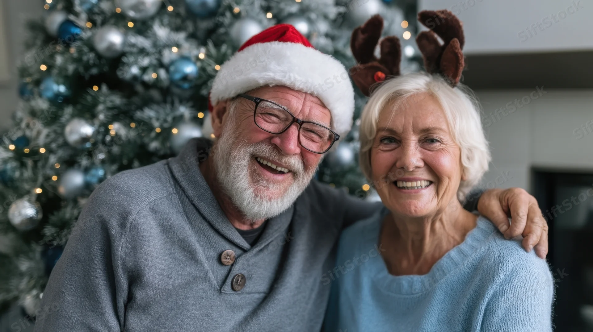 Elderly couple smiling together in front of a decorated christmas tree wearing festive holiday hats