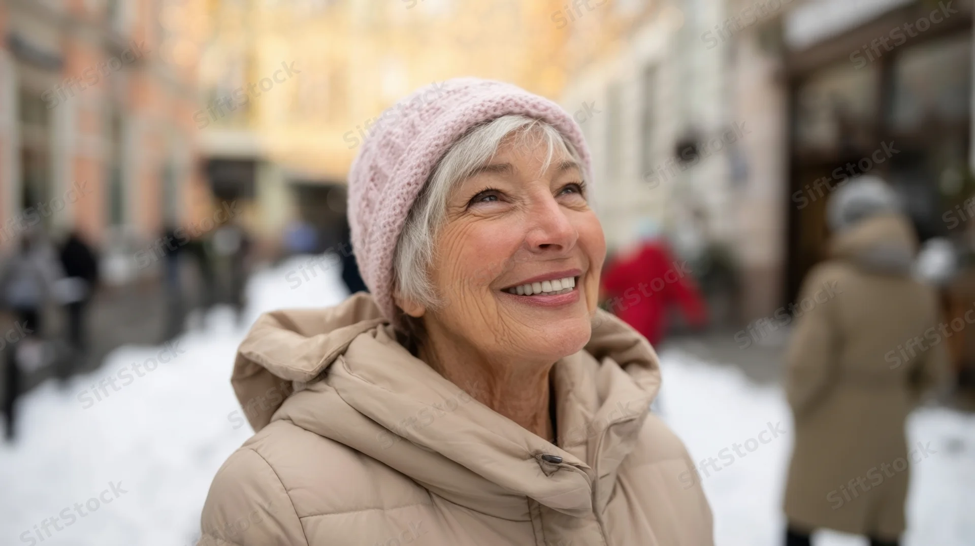 Senior woman smiling outdoors in snowy city street wearing beige coat and pink beanie