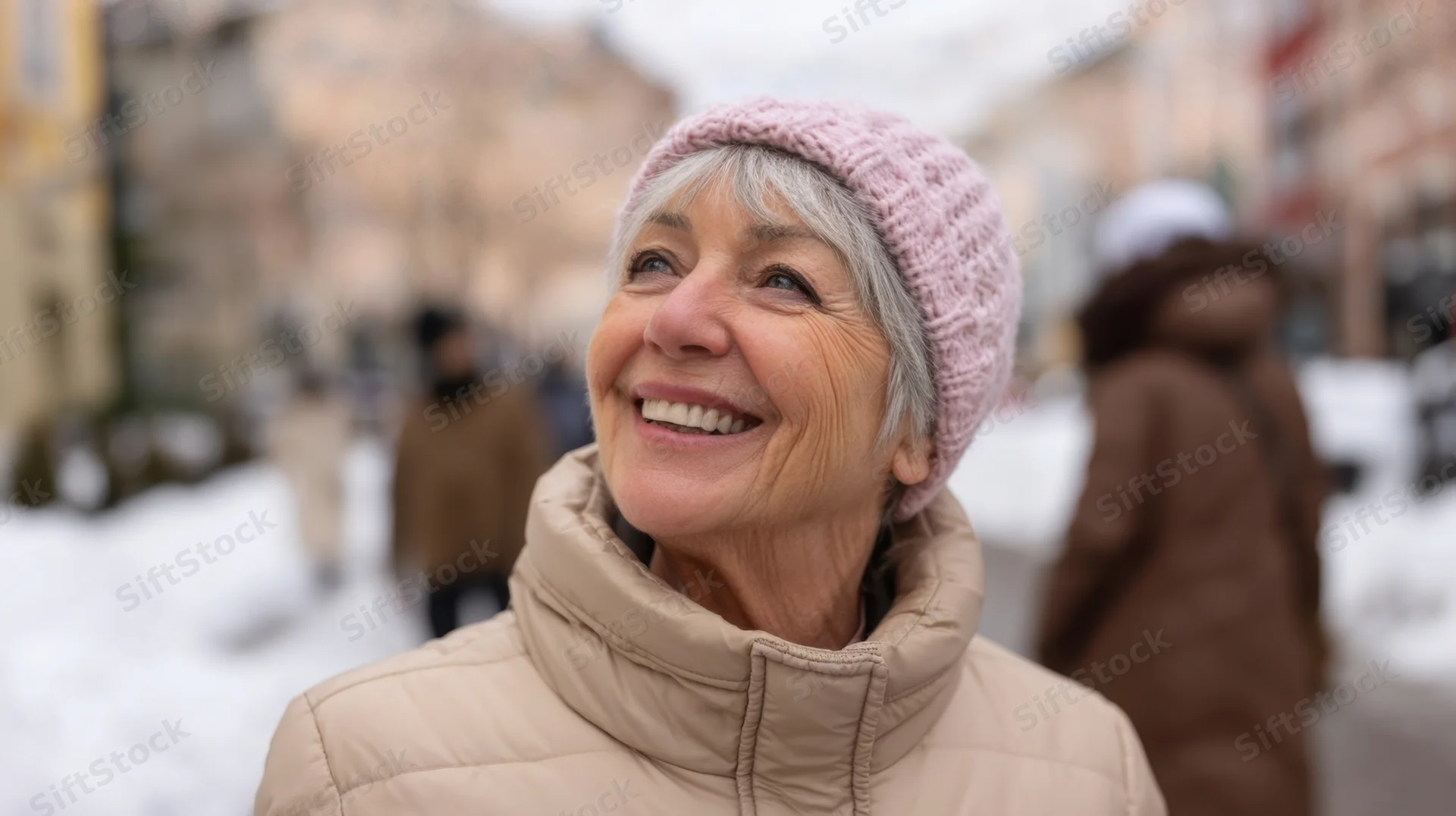 Smiling senior woman in pink beanie and beige coat outdoors in snowy city street