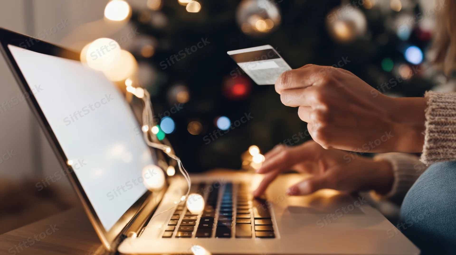 Woman’s hands holding credit card and typing on laptop with christmas tree lights in background